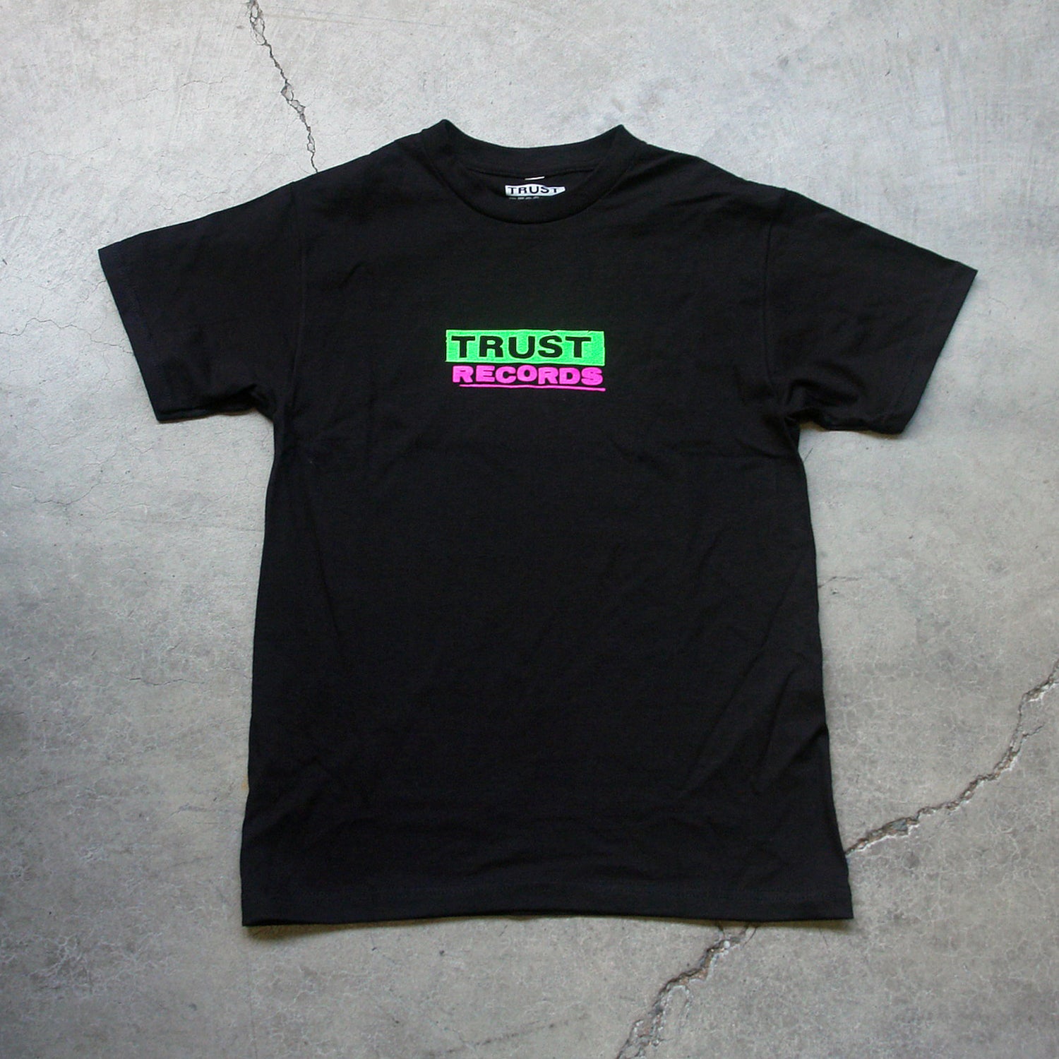 A black Trust Records 1301 Alstyle T-shirt lies flat on concrete. Made of 100% cotton, it features TRUST RECORDS silkscreened on the chest—TRUST in green over pink, RECORDS in pink over green.