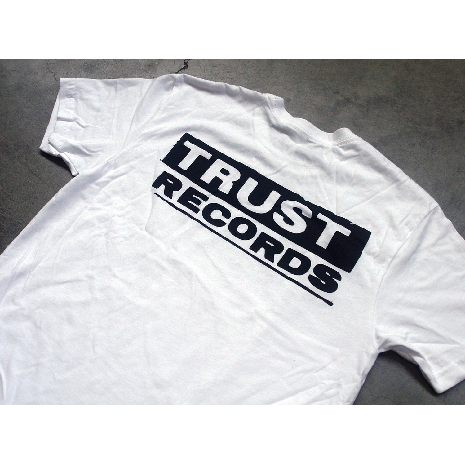 A white Trust Records T-shirt with the bold black logo across the back is displayed on a gray surface.
