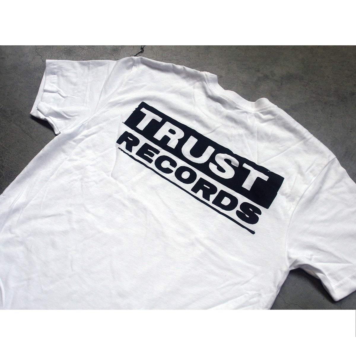 A white Trust Records T-shirt with the bold black logo across the back is displayed on a gray surface.