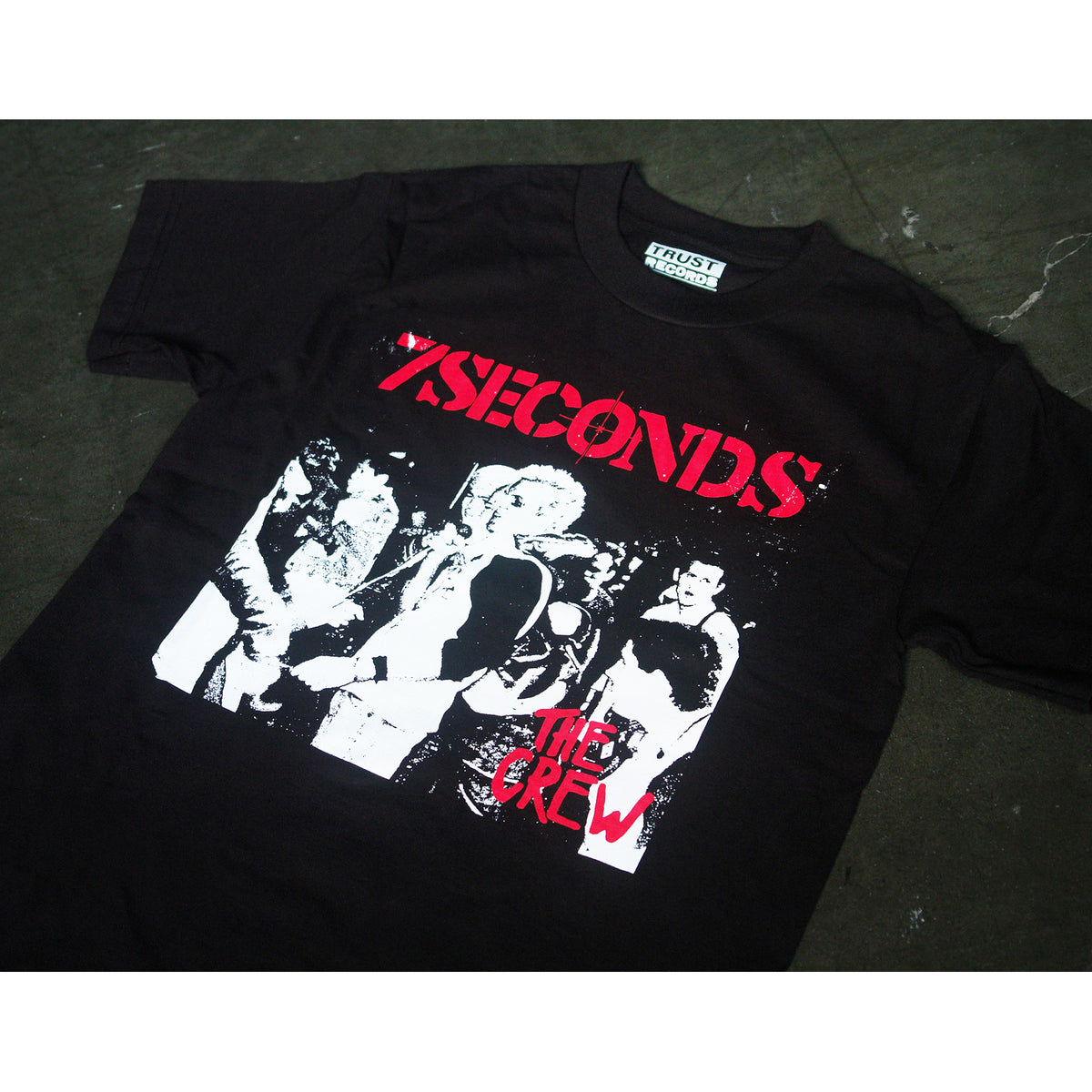 A black 7 Seconds Alstyle 1301 T-shirt features a white and red silkscreened crowd graphic with bold "7SECONDS" in red and "THE CREW" beneath, made from 100% cotton and displayed flat on a dark surface.