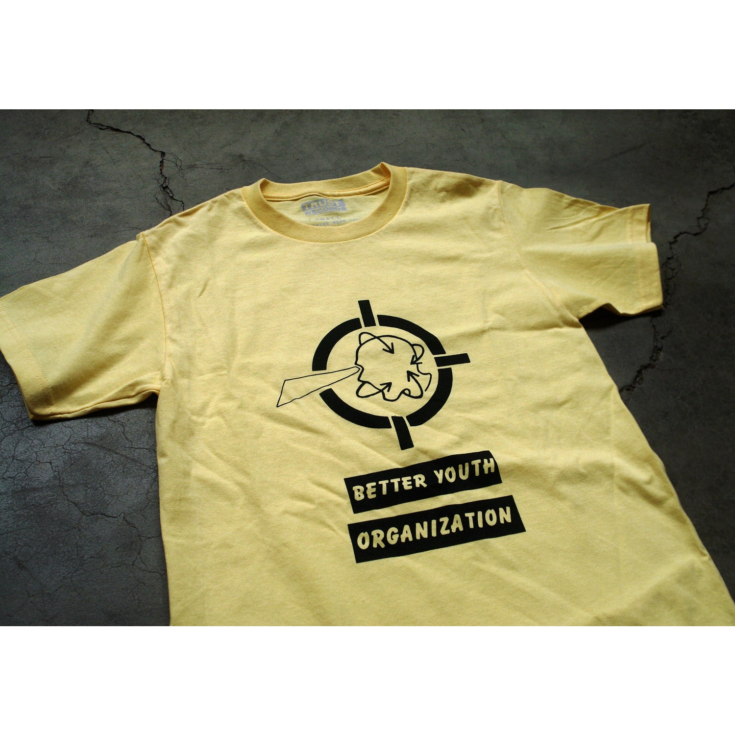 A yellow Better Youth Organization Alstyle 1301 T-shirt rests on gray concrete. It features a black cartoon bird and target silkscreened above BETTER YOUTH ORGANIZATION text. Made from premium 100% cotton.