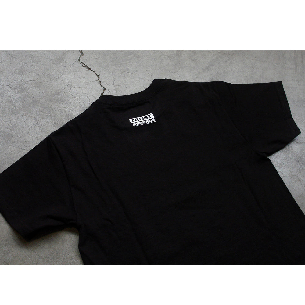 A black 100% cotton Better Youth Organization T-shirt lies on cracked concrete, showing the back with a small white Trust Records label near the neckline.