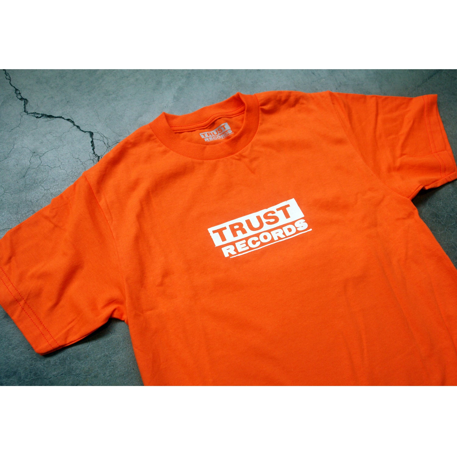 Bright orange Alstyle 1301 T-shirt from Trust Records, featuring bold white "TRUST RECORDS" text silkscreened on the chest, displayed flat on a gray cracked concrete background.