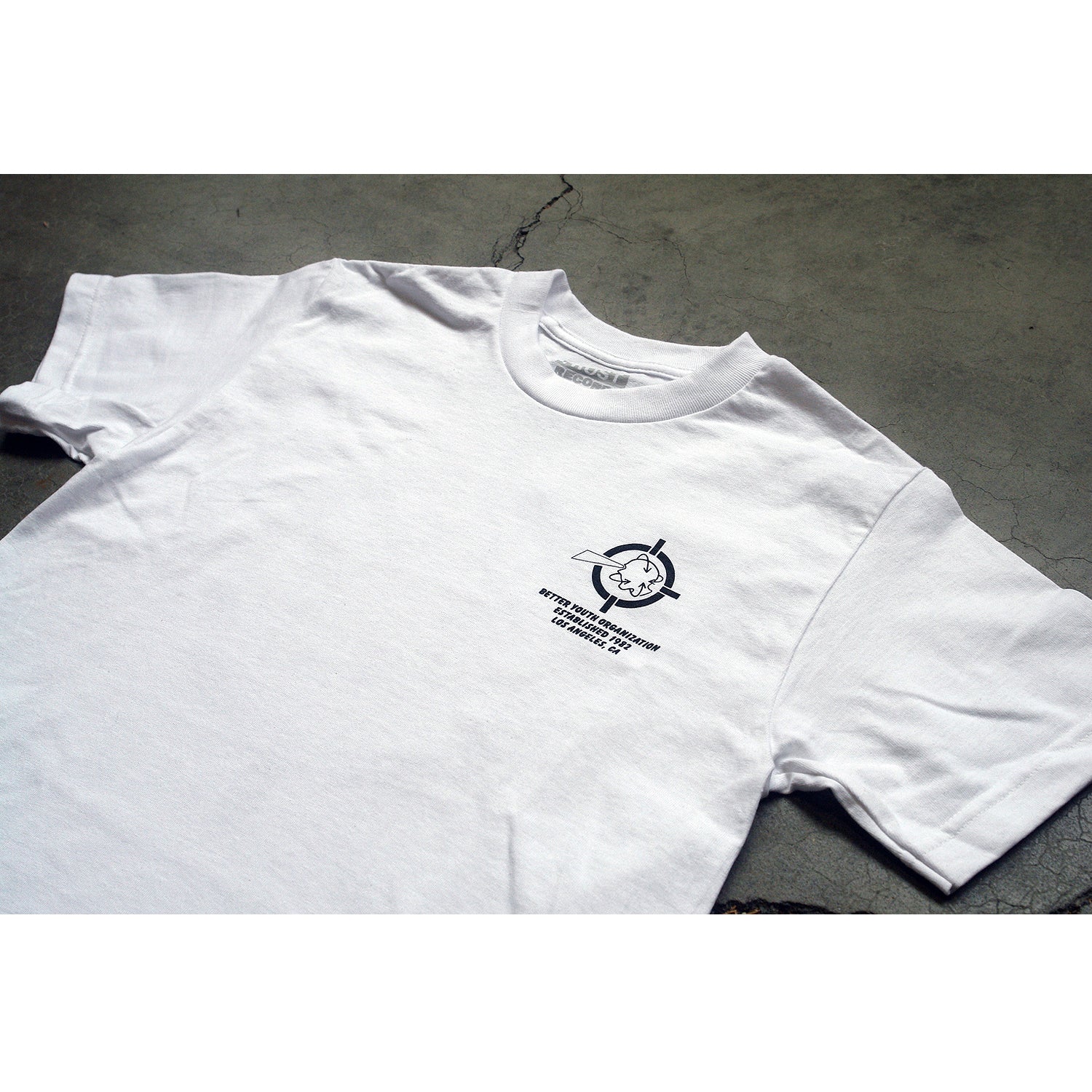 A white Better Youth Organization Alstyle 1301 cotton t-shirt with a small black silkscreened graphic and text on the left chest, featuring a stylized face with circular elements, is laid flat on a gray concrete surface.