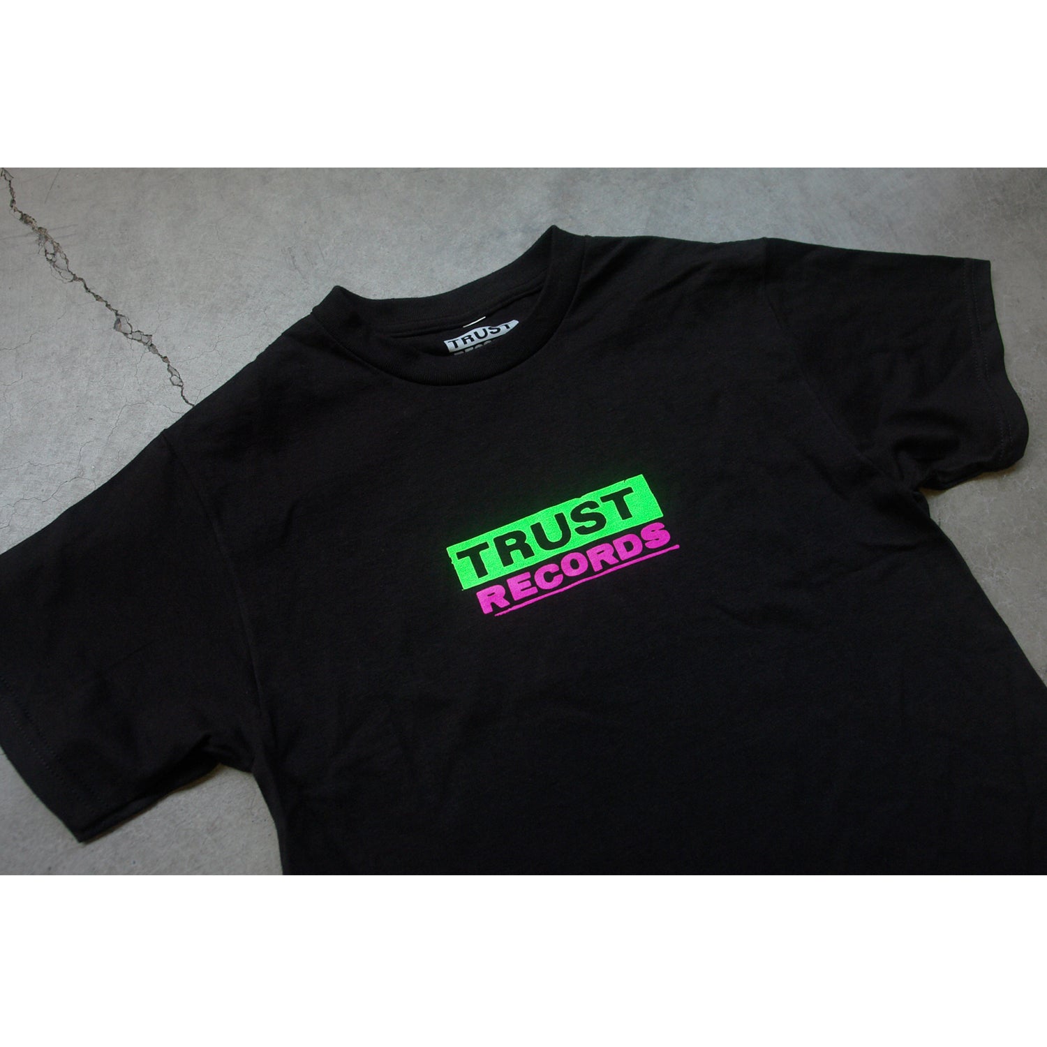 A black Trust Records Alstyle 1301 T-shirt, made of 100% cotton, lies flat on a gray surface with bold silkscreened TRUST in green and RECORDS in pink across the front.