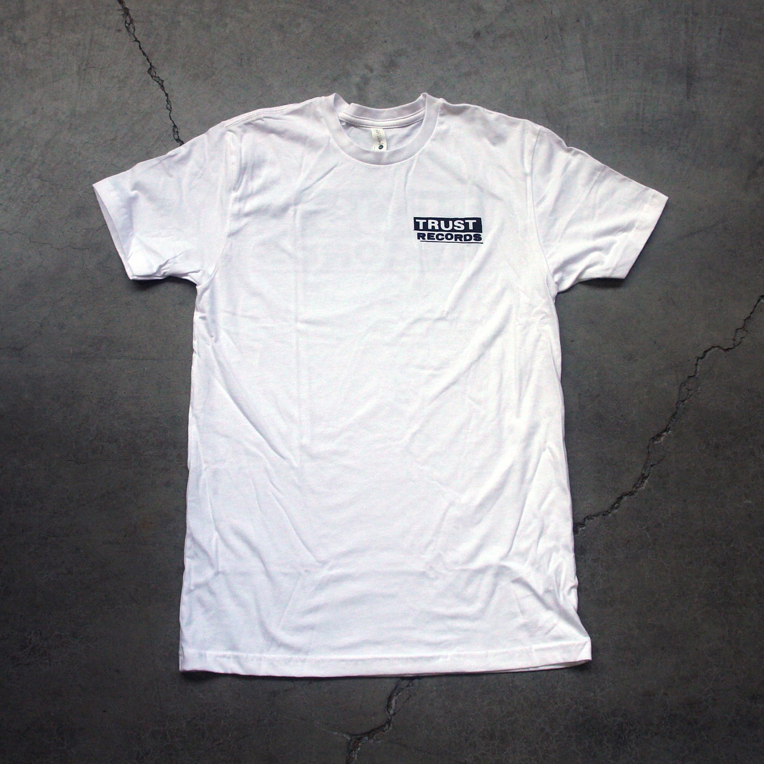 A plain white Trust Records T-shirt lies flat on a gray, cracked surface, featuring a small black and white Trust Records logo on the left chest for a subtle touch of style.