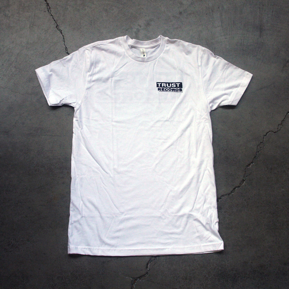 A plain white Trust Records T-shirt lies flat on a gray, cracked surface, featuring a small black and white Trust Records logo on the left chest for a subtle touch of style.