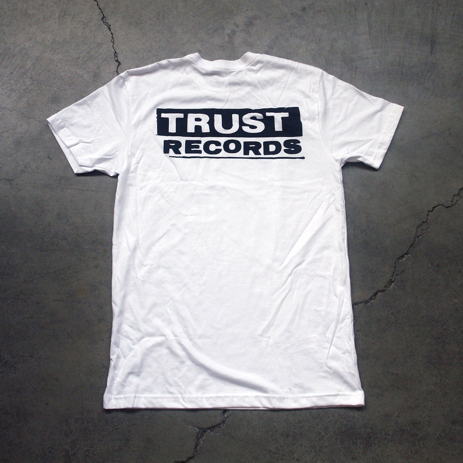 A white Trust Records T-shirt is laid flat on a gray concrete surface, with the bold black Trust Records logo printed on the back.