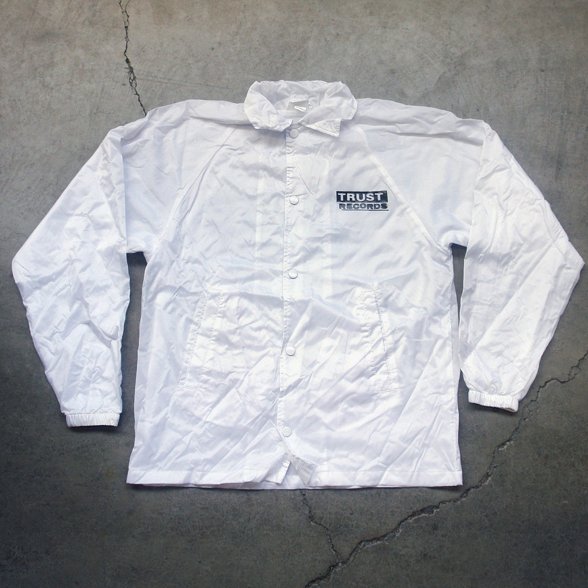 A white Trust Records windbreaker with elastic cuffs and a small logo on the left chest, shown on a gray concrete surface.